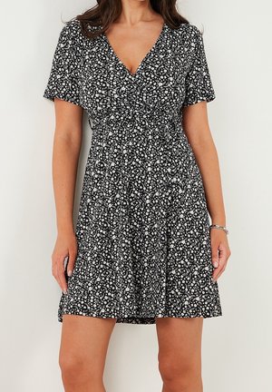Femme portant une robe noire à manches courtes avec un motif floral blanc et un col en V, debout contre un mur clair uni.