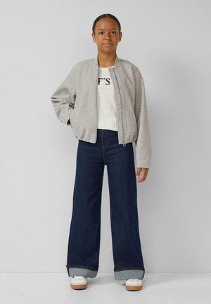 Grijze zip-up bomberjack met een ribgebreide kraag, gecombineerd met een witte top met zwarte opdruk en brede donkerdenim jeans met opgerolde zoom.
