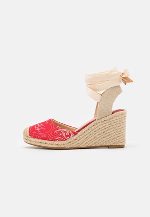 Rød espadrille kile sandal med hvid paisley mønster, beige flettet hæl og cremefarvet ankelbind på hvid baggrund.