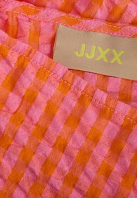Tessuto con un motivo a scacchi in tonalità di rosa e arancione. Presenta un'etichetta con "JJXX" in giallo. Il materiale appare leggero e tessuto.