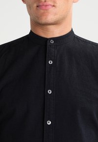 Camisa negra con cuello y parte frontal con botones plateados. La tela parece tener textura con un acabado suave y detalles mínimos.