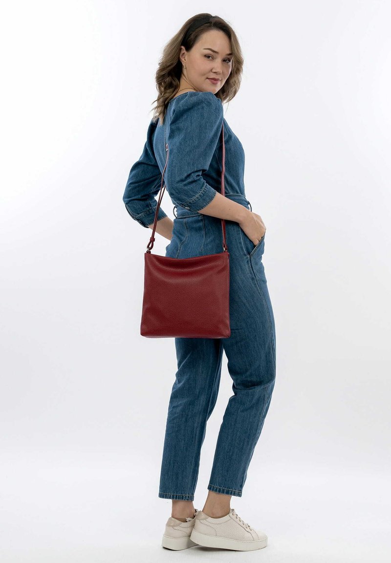 Sac en cuir rouge porté en bandoulière avec une texture lisse, de forme rectangulaire et une longue sangle, porté par une personne en tenue en denim et baskets.