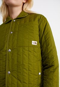 Femme portant une veste matelassée vert olive avec des boutons-pression et le logo The North Face sur la poche poitrine, sur fond blanc.