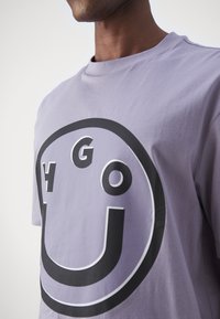 Lilla bomulds t-shirt med et stort sort smilende ansigt i midten af brystet, med fed skrift og en afslappet pasform.