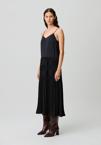Abito nero senza maniche con fondo plissettato e parte superiore liscia. Il modello indossa stivali marroni al ginocchio. Design semplice, senza motivi o accenti.