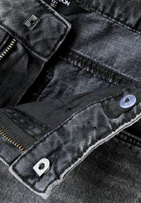 Pantalones de mezclilla negra con cremallera, herrajes plateados y costuras en contraste. Textura suave con un acabado ligeramente deslavado.