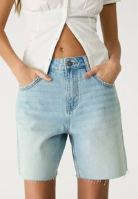 Svetlo modri jeans kratki hlače z ohlapnim fitom, raztrganim roba, petimi žepi in zadrgo. Material kaže subtilno bledenje in teksturo.