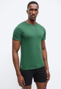Homme portant un t-shirt vert ajusté à manches courtes et un short noir, debout devant un fond clair uni.