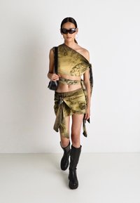 Zweiteiliges Outfit mit einem grün-braun gemusterten Wickeltop und einem Rock mit Knotelement. Kombiniert mit knielangen schwarzen Stiefeln.