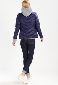 Námořnická prošívaná bunda s chevronovým stehem, šedá mikina s kapucí a žebrované lemy. Kombinováno s tmavými džíny a bílými teniskami, pohled zezadu.