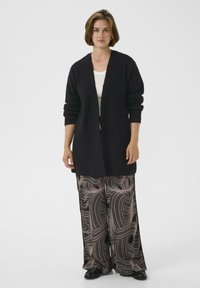 O femeie îmbrăcată cu un cardigan lung negru, un top alb și pantaloni cu pantaloni largi și model, purtând pantofi negri, stând în fața unui fundal alb simplu.