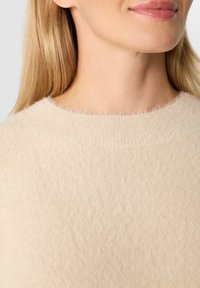 Frau mit heller Haut und langen blonden Haaren, die einen weichen, fusseligen beigen Pullover mit Rundhalsausschnitt trägt, gezeigt von Hals bis zur oberen Brust.