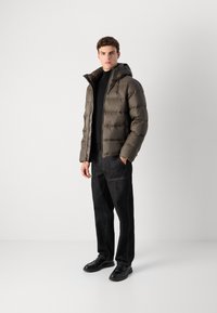 Rutete olivenfarget quiltet pufferjakke med hette, hel glidelås foran og sidelommer, slitt over en svart turtleneck og mørke jeans. Sorte sko.