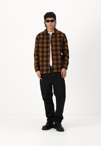 Brun og sort plaid skjorte, knap-op design, parret med sorte bukser og sorte sko. Accessories inkluderer sorte solbriller. Enkel, afslappet stil.