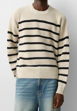 Personne portant un pull en tricot beige avec des rayures horizontales noires et un jean en denim bleu clair sur un fond blanc uni.
