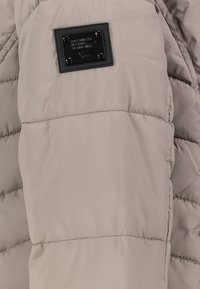 Puffjacke in hellem Beige, mit gesteppter Textur, weichem Synthetikmaterial und einem schwarzen Etikett mit Prägedetails.