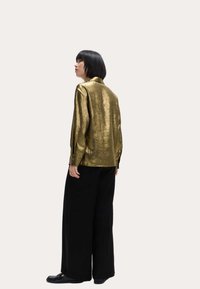 Camisa metálica dorada de manga larga con cuello, pliegues en la parte posterior y puños abotonados, combinada con pantalones anchos negros y zapatos planos negros.