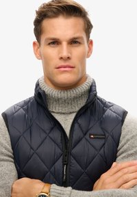 Marineblauwe, gewatteerde puffergilet met hoge kraag, ritssluiting en logo op de borst, gedragen over een lichtgrijze geribbelde trui.
