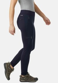 Pantaloni sportivi blu navy con vita alta, tasche laterali con zip e design aderente. Indossati con scarpe da escursione marroni.