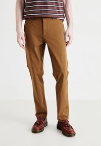 Pantalones de algodón marrón con un diseño de pierna recta, que presentan una cremallera y trabillas para cinturón. El modelo lleva una blusa de rayas y calzado marrón.