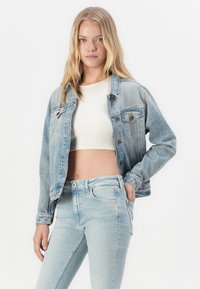 Light blue denim jacket with two chest pockets, button closure, and a relaxed fit, worn over a white crop top and light blue jeans.