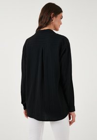 Black long-sleeve shirt featuring a textured fabric, gathered back detail, and buttoned cuffs. Worn with white pants.