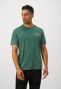 Grøn bomulds T-shirt med rund hals, korte ærmer og et bleget udseende. Har et lille grafisk motiv på venstre bryst.