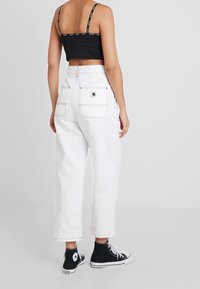 Kvinna som bär vita högmidjade byxor med svarta sömmar och bakfickor, en svart kortärmad topp och svarta högskaftade sneakers.