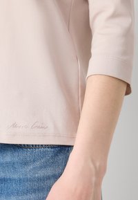Lichtroze, kort mouwtop van zachte stof met een afgeronde zoom en een logo gestikt aan de onderkant, gecombineerd met blauwe denim.