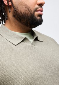 Κοντινό πλάνο του κάτω μέρους του προσώπου ενός άνδρα με γενειάδα και dreadlocks, που φοράει μια υφασμάτινη μπεζ πουκάμισο με γιακά πάνω από ένα ανο ιχτό πράσινο μπλουζάκι.