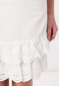 Jupe blanche avec un ourlet à volants superposés, ornée d'une délicate broderie à œillets, réalisée dans un tissu lisse et léger. Aucun autre détail visible.