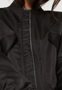 Veste bomber noire avec fermeture éclair à l'avant, détails matelassés et poches plates. Texture lisse avec poignets et taille élastiqués.