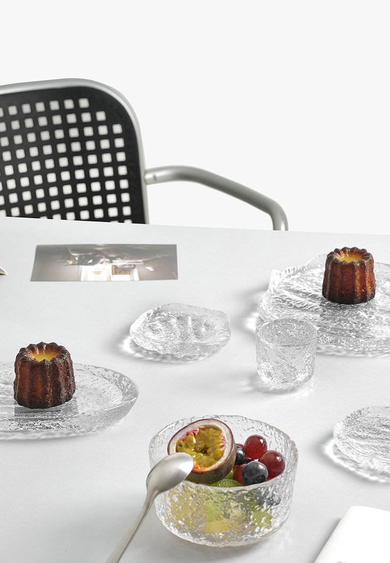 Duidelijk gestructureerd glazen tafelgerei in verschillende vormen; bevat twee cannelés, een assortiment aan fruit en een lepel op een witte tafel.