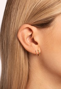 Oreille droite avec deux petites boucles d'oreilles fines en or, cheveux blonds clairs retroussés derrière l'oreille, vue en gros plan.
