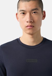Námořnická modrá tričko s kulatým výstřihem vyrobené z hladké tkaniny, s černým vyraženým logem „BOGNER“ na přední straně, s krátkými rukávy a těsným střihem.
