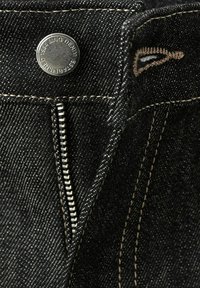 Gros plan sur la fermeture éclair d'un jean en denim noir partiellement ouverte avec un bouton en métal marqué «Establish 1994 IMC Denim» et des surpiqûres beiges.