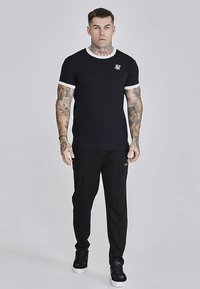 Fekete rövid ujjú póló fehér gallérszegéllyel, kis logóval; fekete nadrággal és fekete tornacipővel párosítva.