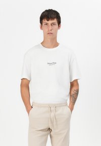 White cotton t-shirt with short sleeves and round neck, featuring a small black logo text on the chest. Paired with beige elastic waist pants.