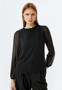 Blusa nera con maniche trasparenti e svasate e scollatura a girocollo. Tessuto morbido con una vestibilità comoda, abbinata a pantaloni su misura rigati.