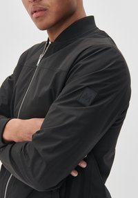 Mustast bomber-jakist, millel on sile pind, soonilised mansetid, kõrge krae ja tekstuurse logoga templiga vasakul varrukal.