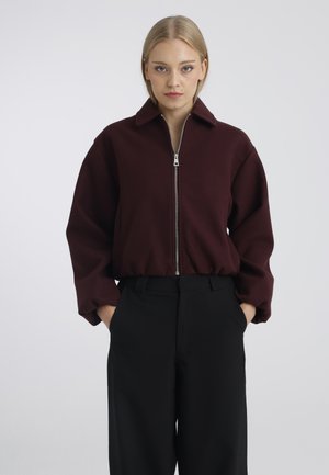 Jeune femme aux cheveux blonds portant une veste zippée bordeaux foncé et un pantalon noir, debout les mains dans les poches devant un fond uni.