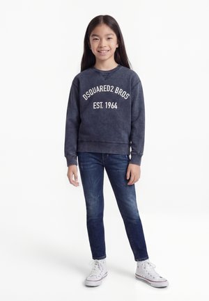 Νεαρή κοπέλα με μακριά σκούρα μαλλιά, φοράει μπλε μπλούζα με λαιμόκοψη, μπλε τζιν, λευκές κάλτσες και λευκά αθλητικά παπούτσια, στέκεται και χαμογελά.