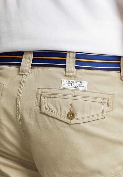 Beżowe chinosy z kwadratową tylną kieszenią, zapinaną na guzik. Powyżej pasa widoczny jest paskowany pas w odcieniach niebieskiego i złotego.