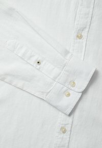 Camisa blanca de manga larga con tela texturada, puño abotonado y dos botones redondos con un ligero brillo. Diseño simple y clásico.