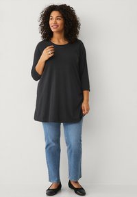 Zwarte tuniek met lange mouwen in een ontspannen pasvorm, gecombineerd met lichtblauwe rechte jeans en zwarte flats. De stof lijkt zacht en glad.