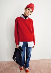 Roter Strickpullover mit Rundhalsausschnitt, über einem weißen Hemd getragen. Dunkelblaue Jeans, orangefarbene Sneakers mit bunten Schnürsenkeln und eine schwarze Tragetasche.