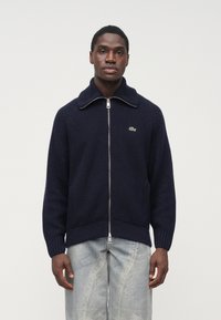 Tmavomodrý pletený sveter na zips s veľkým golierom a rebrovanými detailmi, s logom v zelenej farbe na ľavej strane hrudníka. Skombinovaný so svetlými rifľovými nohavicami.