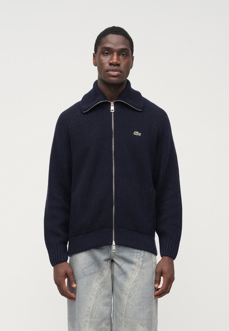 Tmavomodrý pletený sveter na zips s veľkým golierom a rebrovanými detailmi, s logom v zelenej farbe na ľavej strane hrudníka. Skombinovaný so svetlými rifľovými nohavicami.