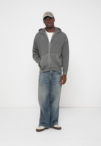 Grauer Reißverschluss-Hoodie mit vorderen Taschen, kombiniert mit lockeren blauen Jeans und weißen Schuhen. Zubehör besteht aus einer hellgrauen Mütze.