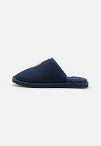 Marineblå slip-on tøfler med en myk, teksturert innvendig og en spiss tådesign. Har detaljert logo sydd på oversiden.